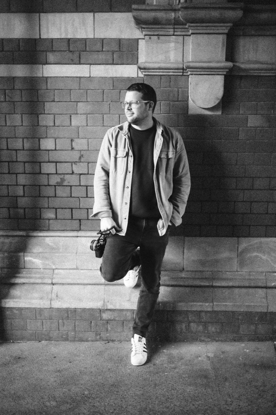 Che leaning against a brick wall with a camera in hand